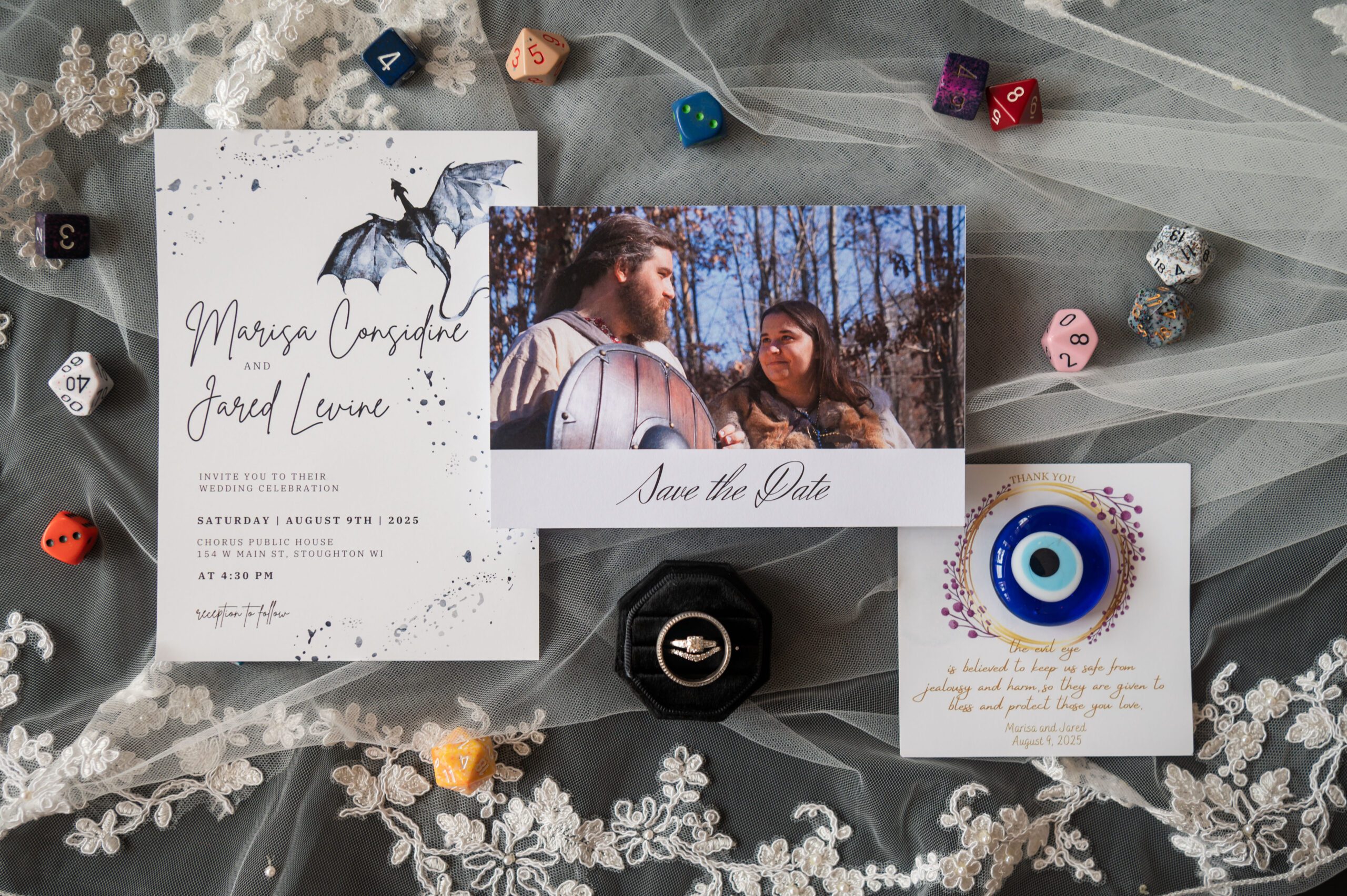 Flat lay of Marissa and Jared’s Dungeons & Dragons inspired wedding details with custom dragon invitation, save the date, evil eye favor, and polyhedral dice at Chorus Public House, photographed by Danielle Lund Photography.