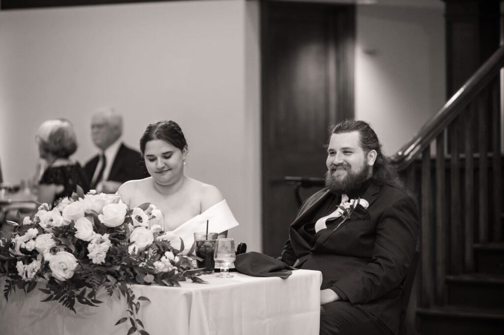 Marissa and Jared seated at their sweetheart table decorated with white roses and greenery during toasts at Chorus Public House wedding reception.